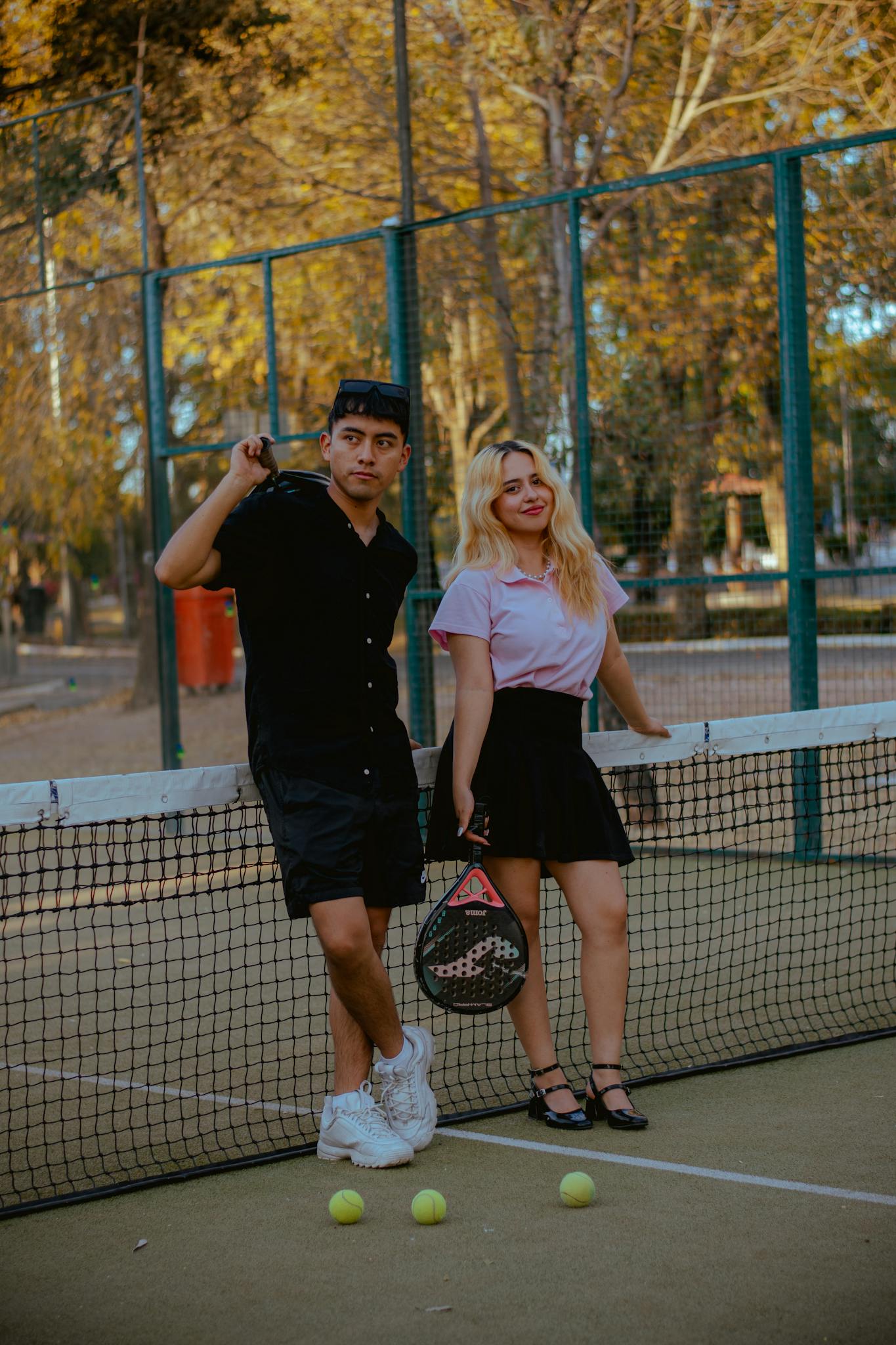 Two young adults leaning against a tennis net, enjoying a sunny day outdoors with rackets and balls in view.